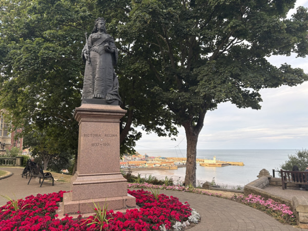 Statue of Queen Victoria on a granite pedestal surrounded by flower beds, with Scarborough’s seafront and townscape visible in the background.