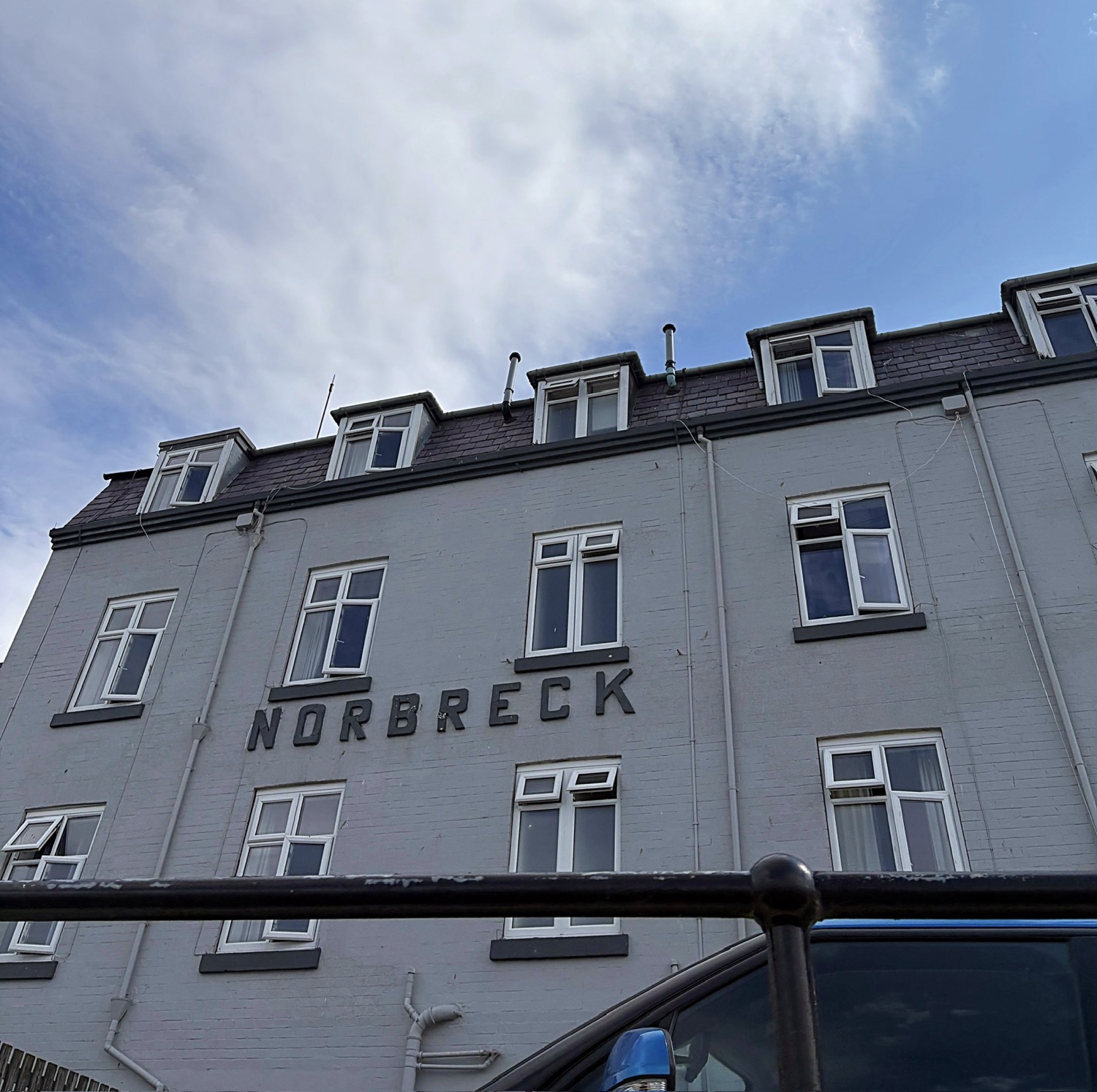 Norbreck Hotel exterior — Front view of the Norbreck Hotel with grey brick façade, white‑framed windows and dormer roofline under a bright, partly cloudy sky.